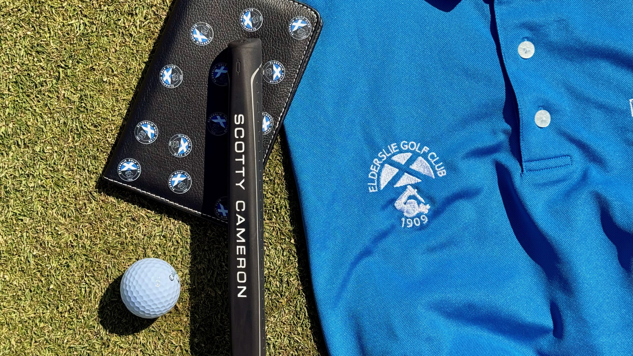 Black Scotty Cameron putter with BMW logo ball markers lying next to blue Elderslie Golf Club polo shirt and white golf ball on artificial grass