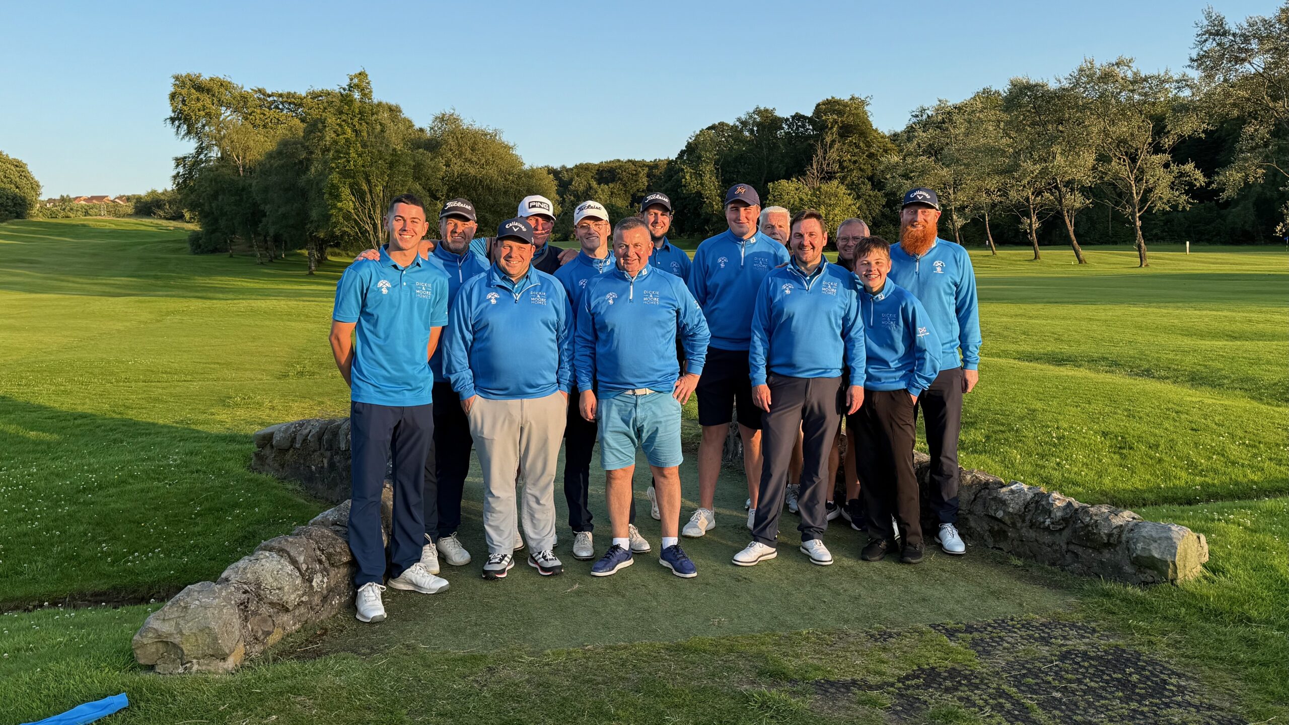 An all male golf team stands on a birdge with the golf course in the background and blue skies overhead